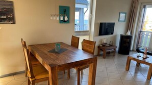 A dining area with a wooden table and chairs, a small side table with a bottle and candle, a TV on the wall, and a view of the sea through a window. - Apartment in Rechlin by Muritz National Park (Rechlin)
