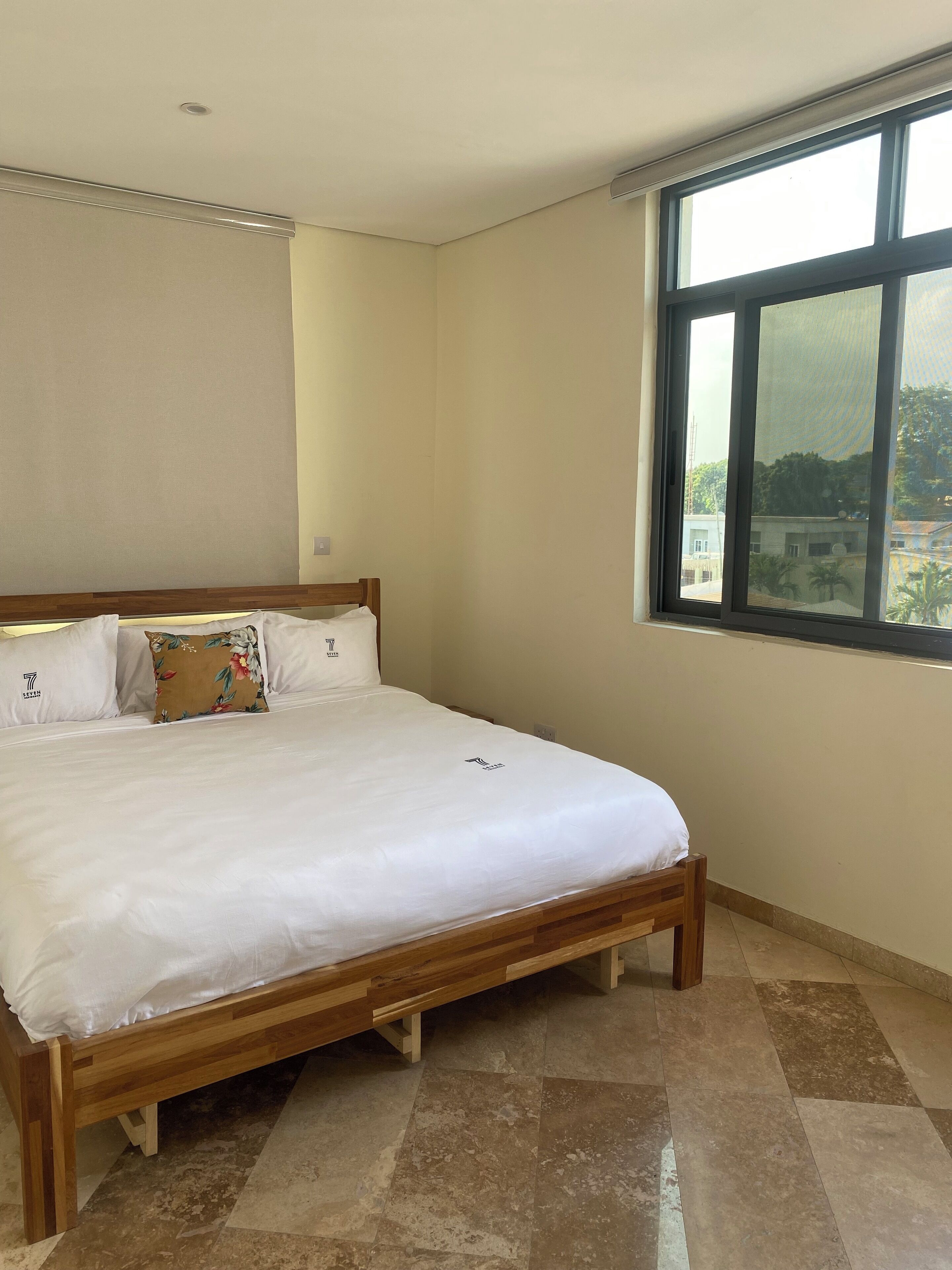 A bedroom with a wooden bed, white bedding, and a large window offering a view of buildings and greenery.