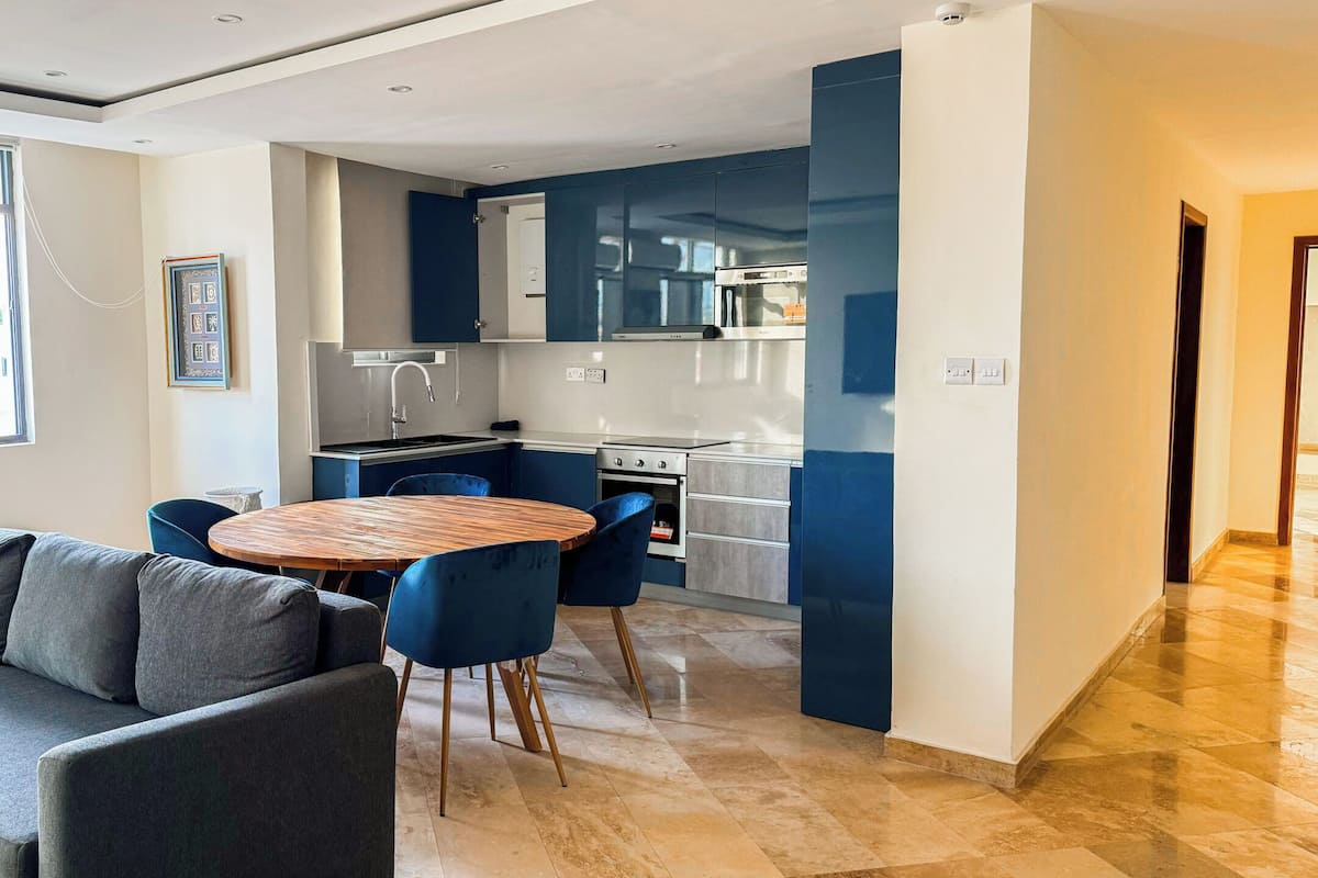 Une cuisine moderne avec des meubles bleus, un coin repas doté d’une table ronde en bois et de chaises bleues, et un canapé gris.
