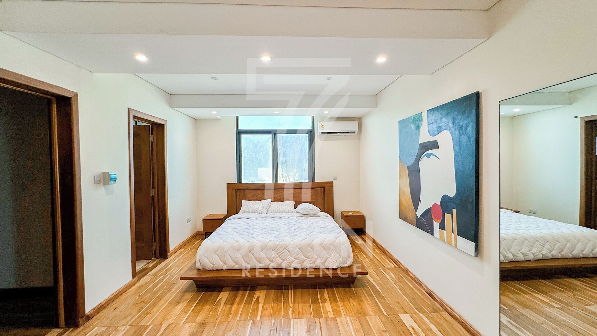 Een slaapkamer met een houten vloer, een bed met wit beddengoed, een nachtkastje, een wandairconditioner en een grote spiegel die de kamer weerspiegelt.