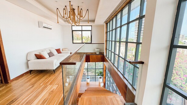 Un intérieur moderne avec un escalier en bois, une balustrade en verre, un canapé blanc agrémenté de coussins et de grandes fenêtres offrant une vue sur l’extérieur.