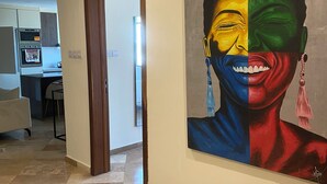 Un couloir avec un sol carrelé, un tableau accroché au mur et un coin cuisine visible à travers une porte ouverte.