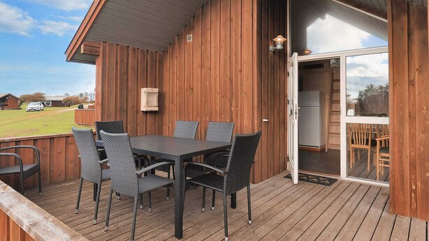 Una terraza de madera con una mesa y sillas, un ventanal corredizo que da a una cocina, y vista a un área verde con un coche al fondo.