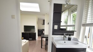 Un baño moderno con un espejo grande, un lavamanos blanco y un mueble de tocador oscuro. Hay un televisor en la zona de estar contigua.