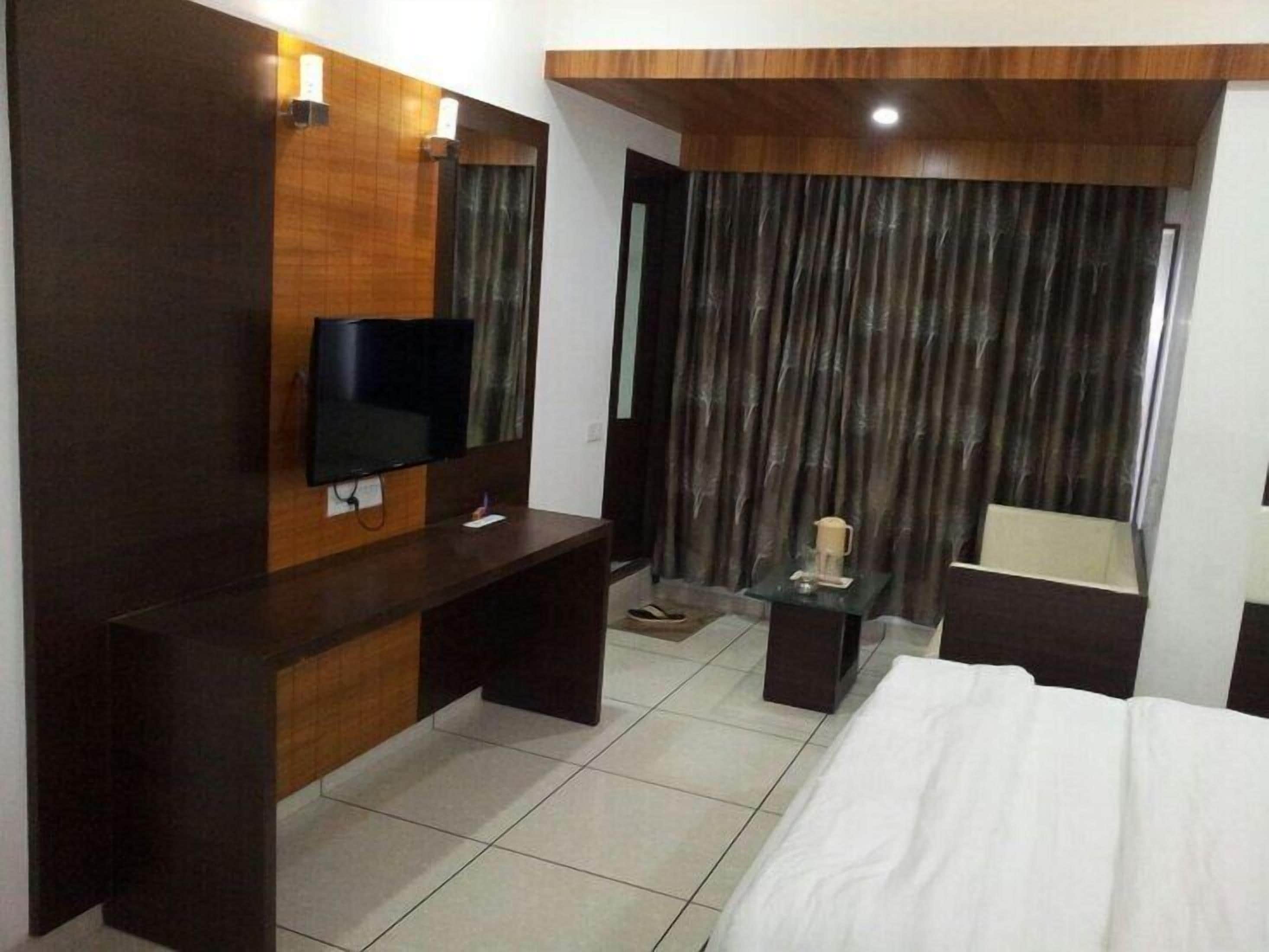 Una habitación de hotel moderna con una cama grande, un televisor de pantalla plana montado en un panel de madera, una mesita de noche pequeña con una lámpara y un telón oscuro de fondo.