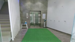 A hallway with a green carpet, a staircase, a glass elevator, and a small table with a spray bottle. - Hotel Apex Inn (Vapi)