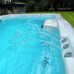 Una piscina amb aigua cristal·lina, envoltada d'una zona verda i un edifici amb terrassa coberta.