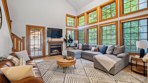 A spacious living room with a fireplace, large windows, a sectional sofa, and a wooden coffee table. - 376 Disciples Village | Game Room | Fireplace | Walking Distance to Slopes! (Boyne Falls)