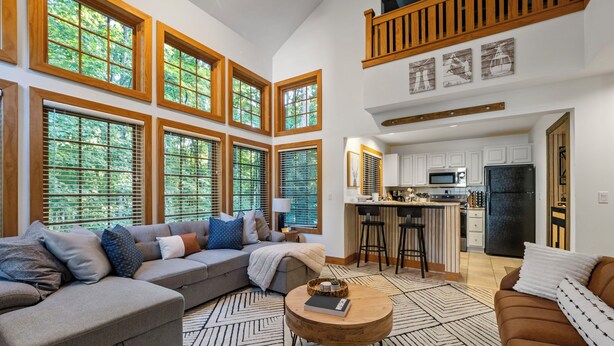 A spacious living room with a large sectional sofa, a coffee table, and a view of the kitchen through large windows. - 376 Disciples Village | Game Room | Fireplace | Walking Distance to Slopes! (Boyne Falls)
