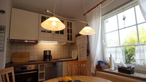 A kitchen with white cabinets, a wooden table with a placemat, a coffee maker, and a window with curtains. - Doppelhaushalfte in Nessmersiel (Dornum)