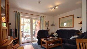 A living room with a sofa, armchair, coffee table, and a television. There is a sliding glass door leading to an outdoor area. - Doppelhaushalfte in Nessmersiel (Dornum)
