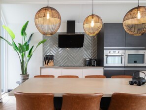 A modern kitchen with a herringbone tile backsplash, a flat-screen TV, stainless steel appliances, and a dining area with a large table and brown chairs.