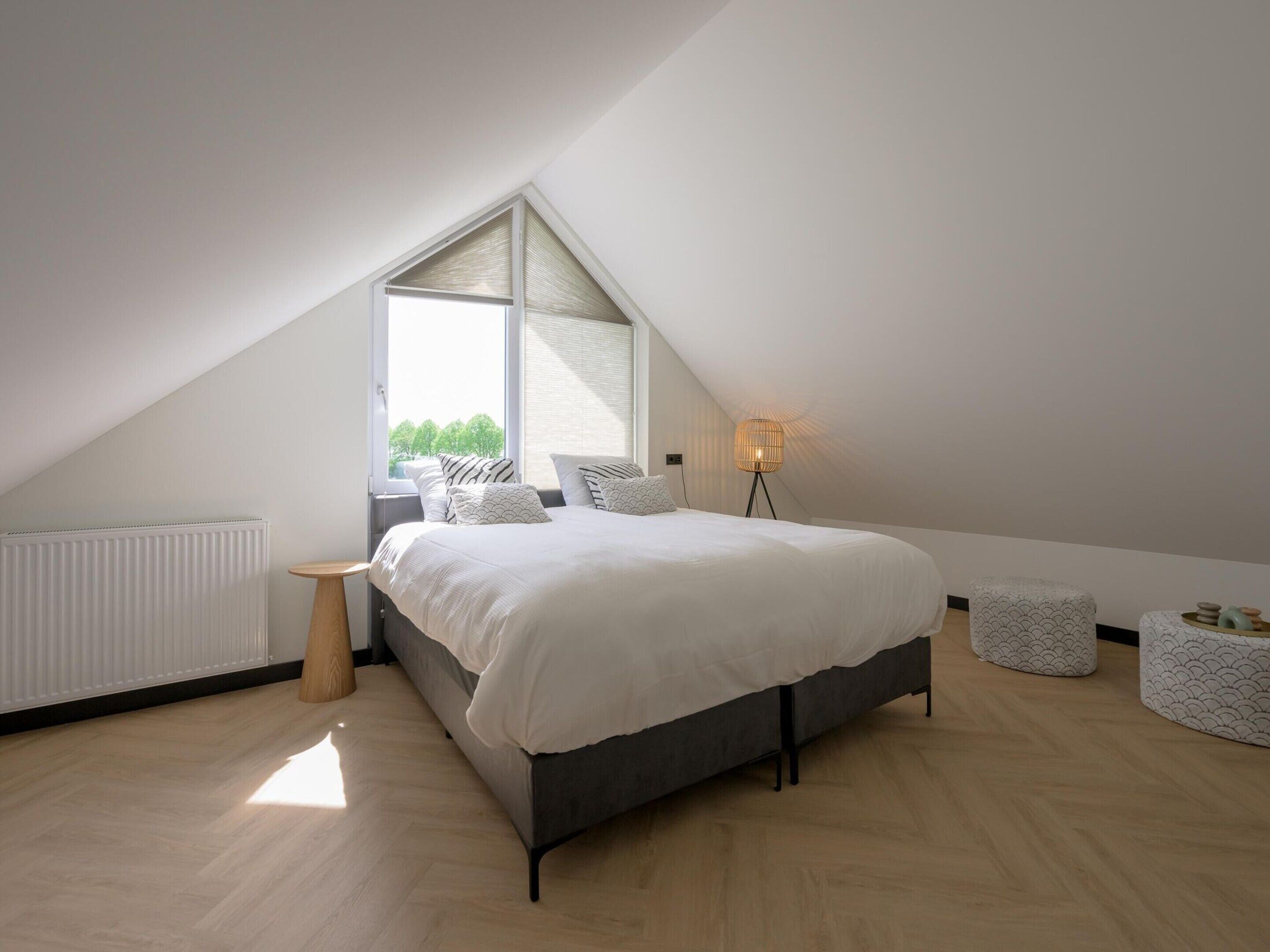 Ein Schlafzimmer mit schrägen Wänden, einem Bett mit grauem Gestell, einem Nachttisch, einer Stehlampe und einem Fenster mit Blick auf Bäume.