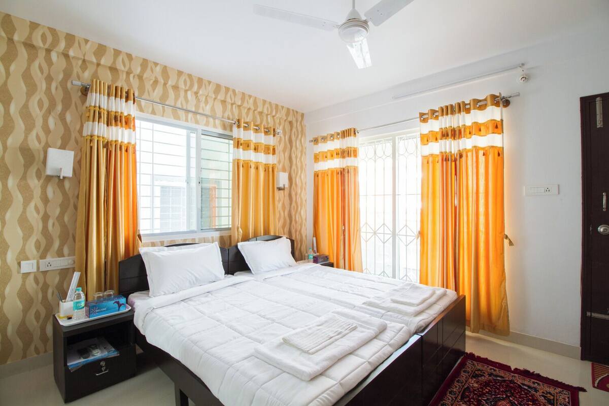 A hotel room with a large bed, two bedside tables, a nightstand, a ceiling fan, and a window with curtains.