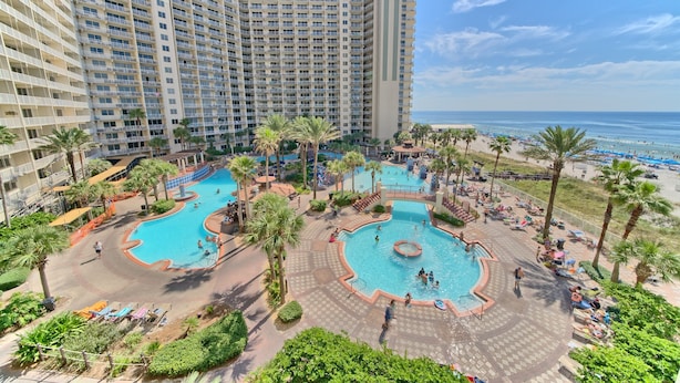 A large hotel with multiple swimming pools, palm trees, and a beach view.