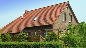 Une maison à deux étages avec un toit recouvert de tuiles rouges, une terrasse en bois et une haie bien entretenue à l’avant.