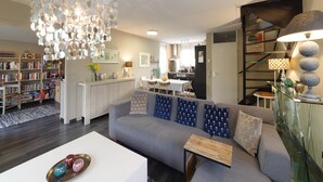 Una sala de estar moderna con un sofá gris, una mesa de centro blanca y una lámpara de techo original. Se observa una estantería llena de libros y una zona de cocina al fondo.