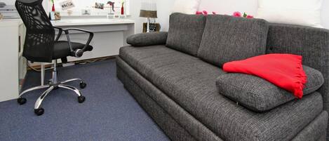 A room with a grey sofa, a black office chair, a desk, and a window with pictures on the wall.