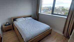 A bedroom with a single bed, a wooden nightstand, a window with curtains, and a view of buildings outside.