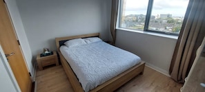 Un dormitorio con una cama individual, una mesita de noche de madera, una ventana con cortinas y vistas a edificios.