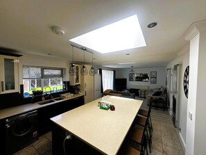 Una cocina moderna con una isla central, un comedor con una mesa alta y un espacio de sala con sofá y televisor.