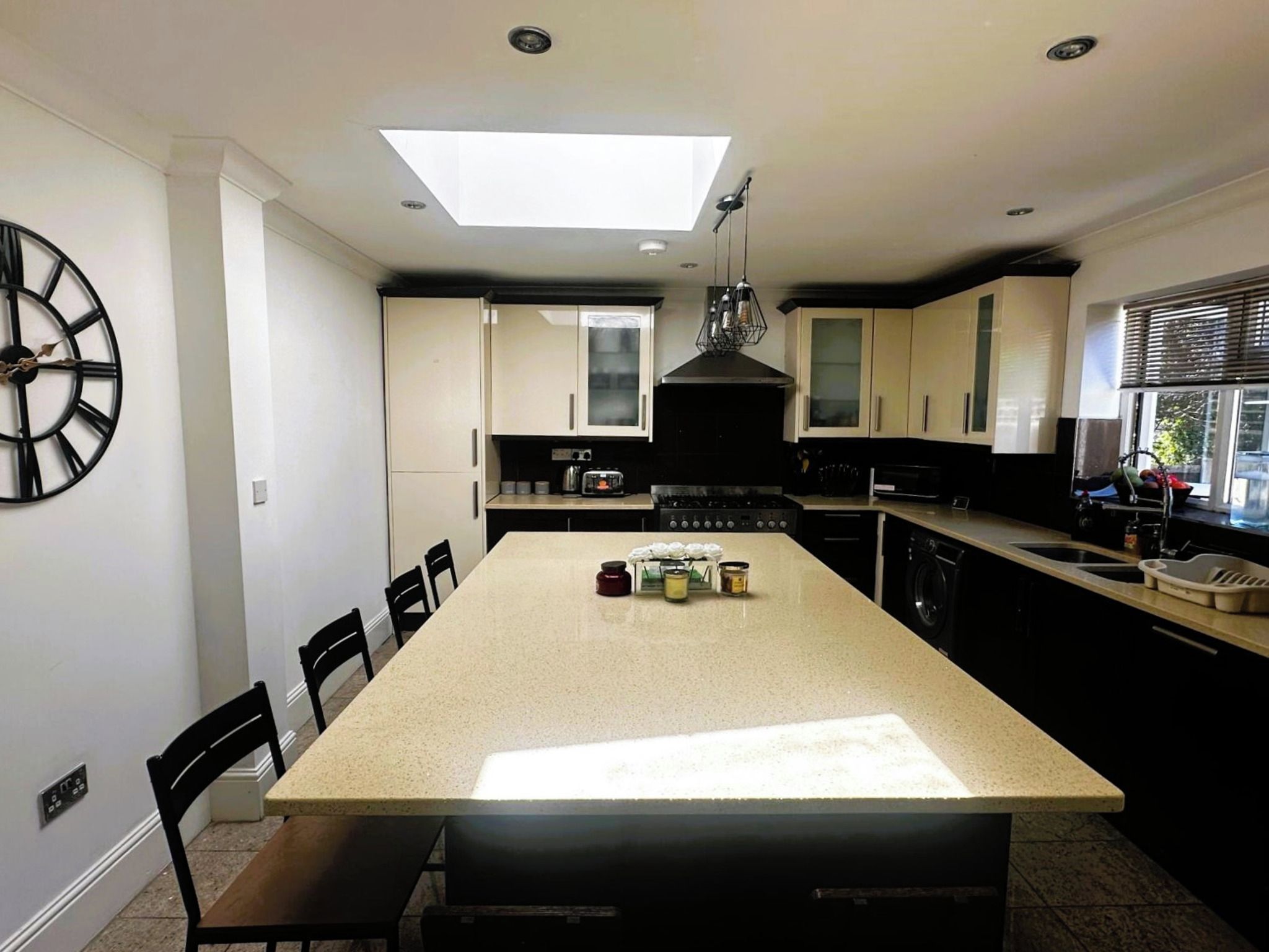 A modern kitchen with a central island, black chairs, a large clock, and a skylight.
