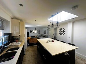 Una cocina moderna con una isla central, un comedor con un reloj grande, y una sala de estar con sofá y mesa de centro.