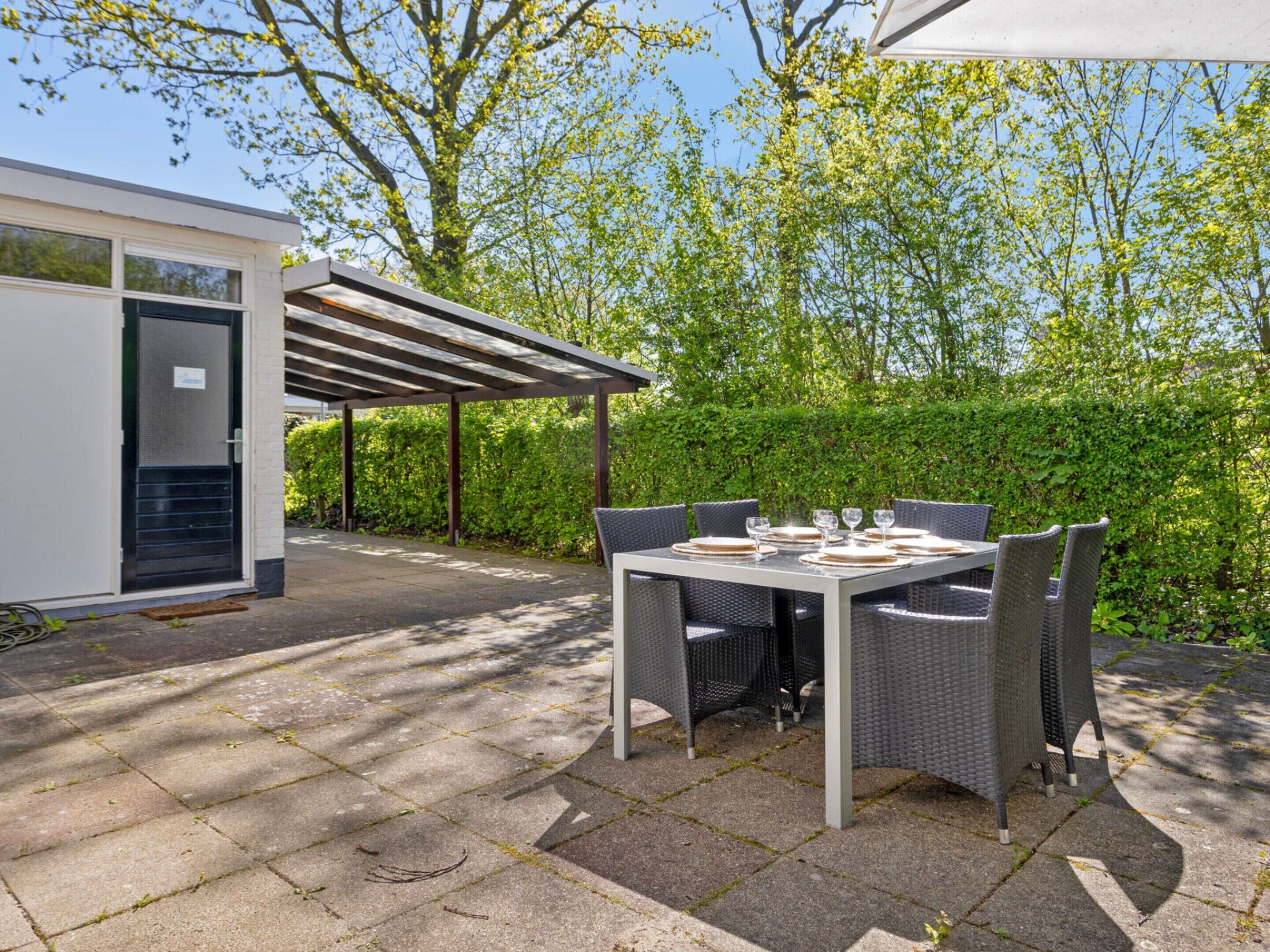 An outdoor patio with a table set for dining, surrounded by greenery and a building with a covered area.