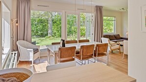 Ett modernt matområde med ett stort bord och stolar, stora fönster med gardiner och utsikt över grönområden utanför.