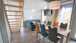 A modern living room with a wooden staircase, a blue sofa, a dining table with chairs, and a TV mounted on the wall. - Ferienhaus im Ferienpark Mirow (Mirow)