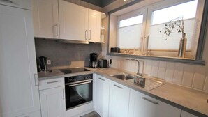 A modern kitchen with white cabinets, a built-in oven, a coffee maker, and a window with blinds. - Ferienhaus im Ferienpark Mirow (Mirow)