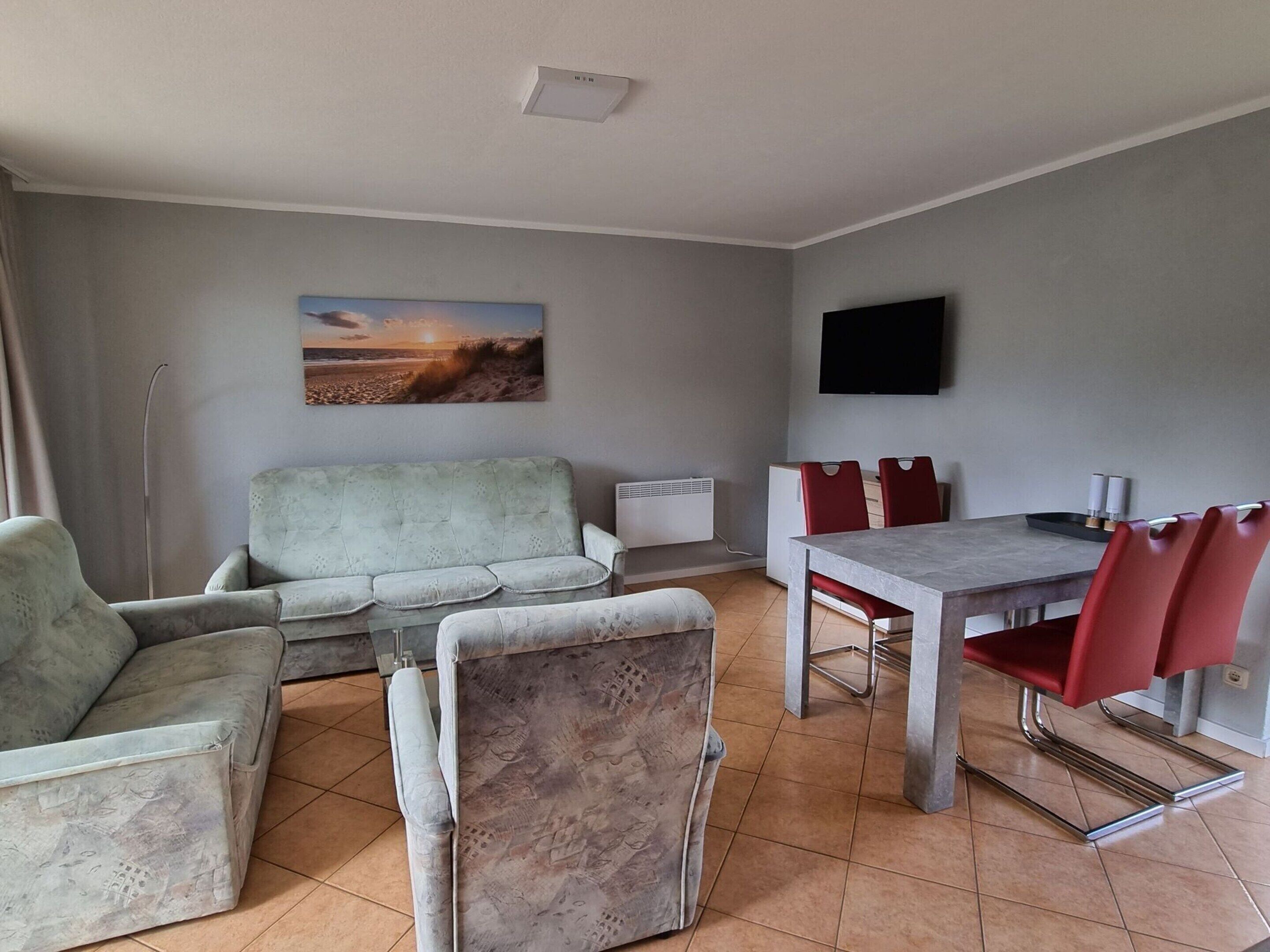 A living room with a grey sofa, a dining table with red chairs, a flat-screen TV, and a painting of a sunset.