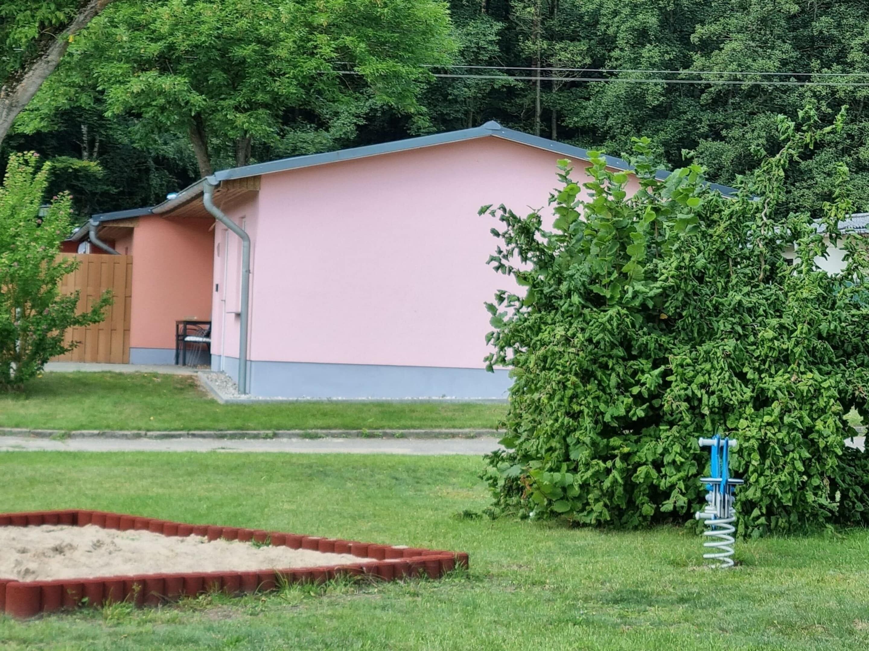 A single-story building with a pink exterior and a blue roof, surrounded by a well-maintained lawn and a dense green bush.