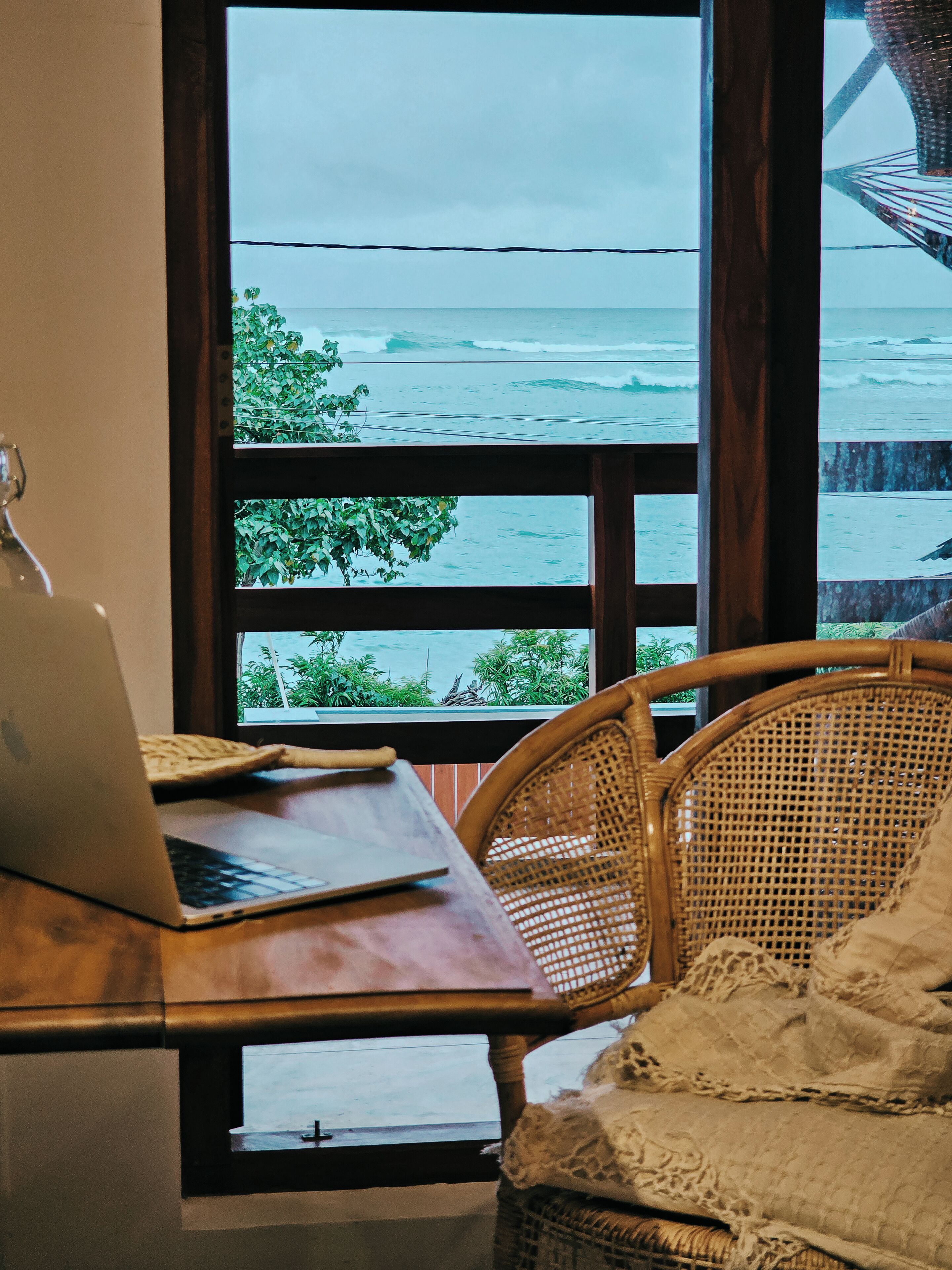 A room with a laptop on a desk, a wicker chair, and a view of the ocean through a window.