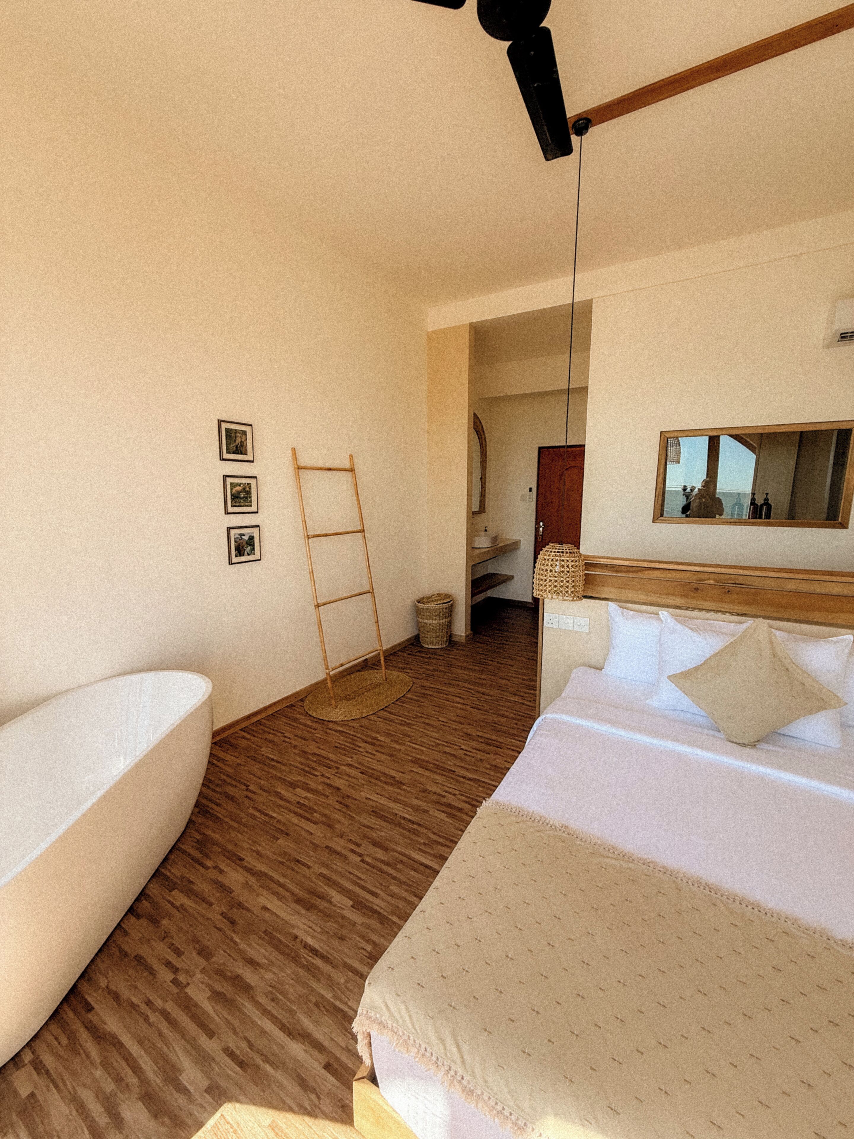 Una camera d'albergo con una vasca da bagno grande, un letto con lenzuola bianche, un pavimento in legno, uno specchio e una trave in legno a soffitto.