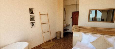 A hotel room with a large bathtub, a bed with white bedding, a wooden floor, a mirror, and a wooden ceiling beam.