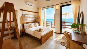Een slaapkamer met een houten bed, een schilderij, een plafondventilator, een groot raam met uitzicht op de oceaan en een houten tafel met stoelen.