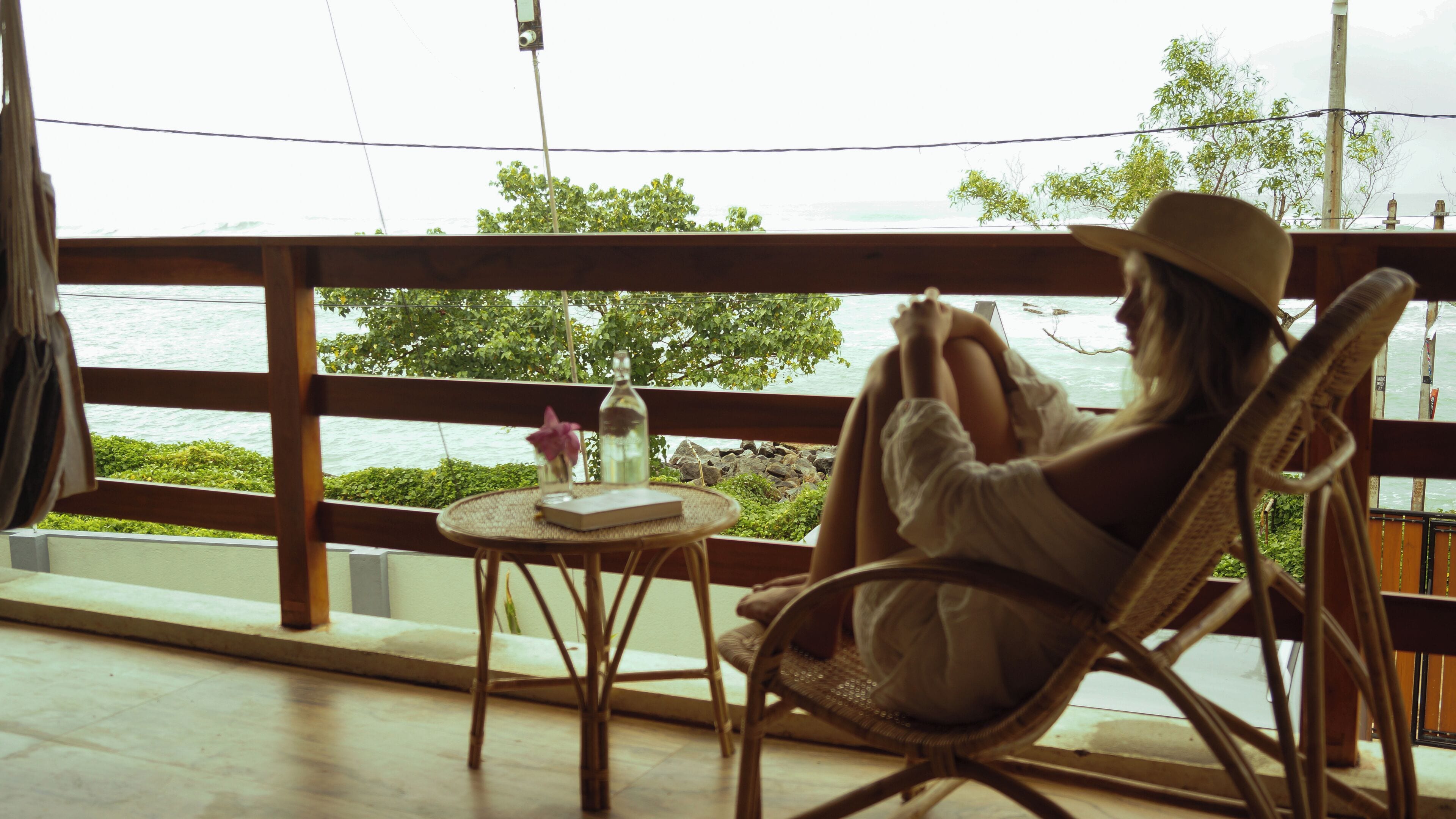 A person sitting on a wicker chair by a window overlooking a body of water, with a small table and a bottle of water in front of them.