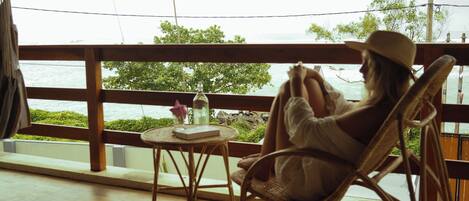 A person sitting on a wicker chair by a window overlooking a body of water, with a small table and a bottle of water in front of them.