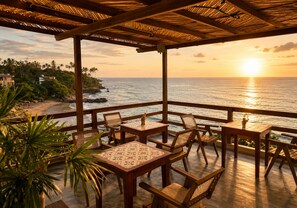 Zona salotto esterna coperta con tavoli e sedie, affacciata su una spiaggia e l'oceano al tramonto.