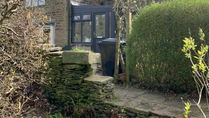 Una casa de piedra con una puerta oscura, una chimenea y un jardín con un muro de piedra y un contenedor negro grande.