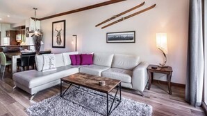 A modern living room with a sectional sofa, a wooden coffee table, and a fireplace. There is a dining area in the background with a chandelier.