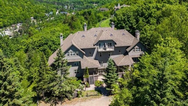 A large, multi-story house surrounded by dense forest, with a mountainous backdrop.