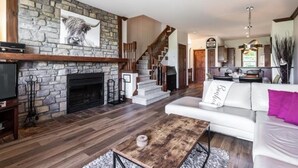 A spacious living area with a stone fireplace, a staircase leading to an upper level, and a modern kitchen in the background.
