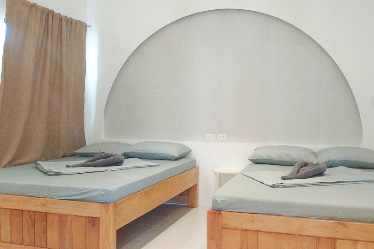 Dos camas de madera con ropa de cama y almohadas grises, una mesita de noche blanca y un amplio ventanal de arco.