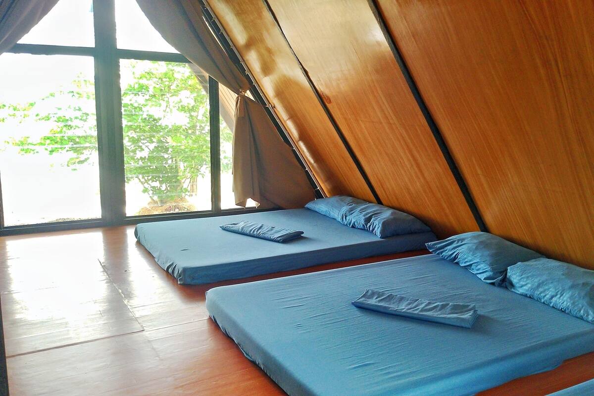 Habitación con dos camas, cada una con colchón y almohadas azules, suelo de madera y un ventanal con cortinas.