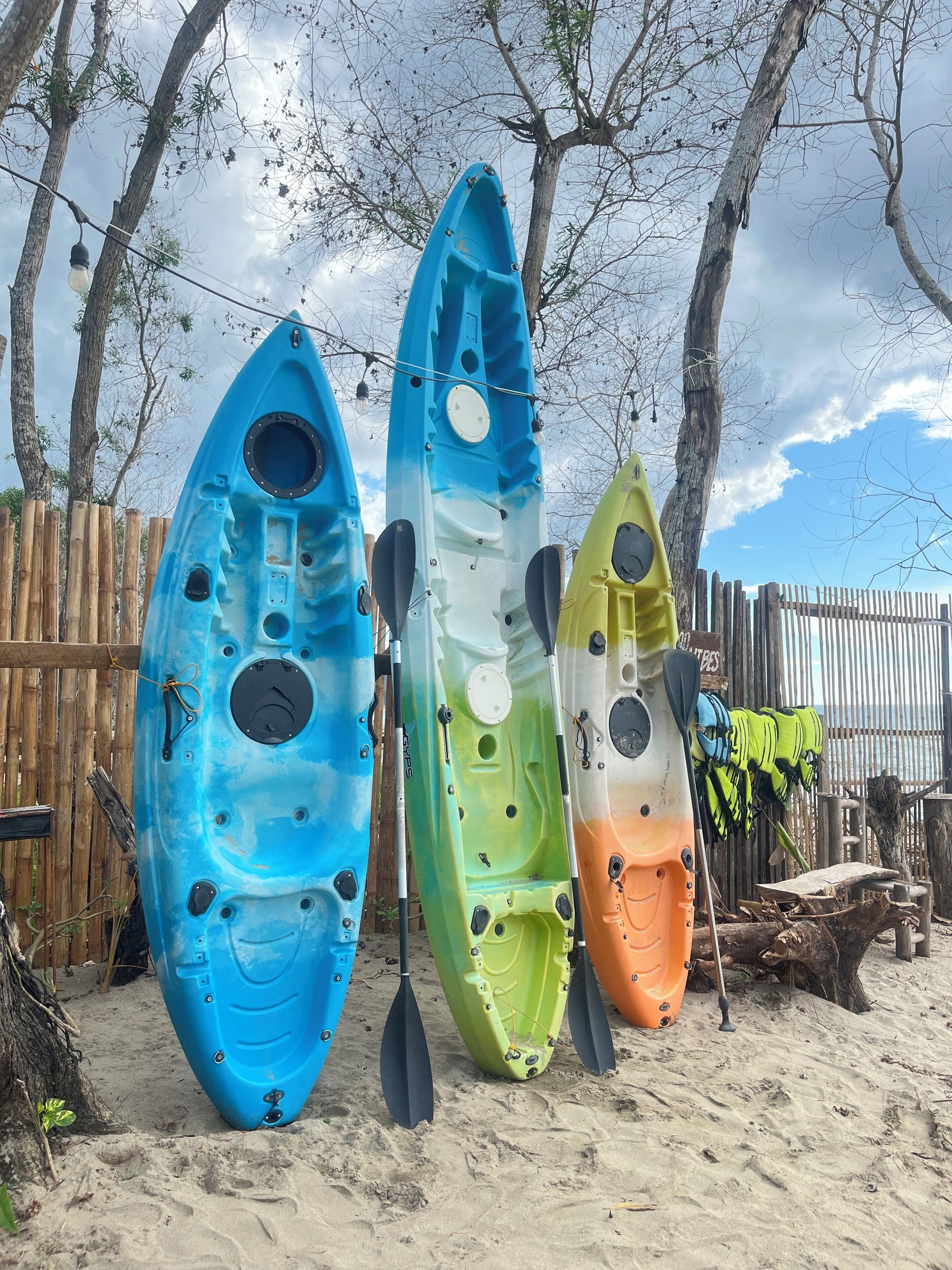 Tres kayaks alineados en una playa arenosa, con una valla de madera y árboles al fondo.