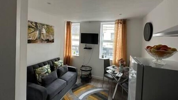 A compact living space with a sofa, a flat-screen TV, a dining area with a table and chairs, and a refrigerator. - FC Platinum Accommodation (Watford)