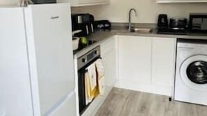 A compact kitchen with white cabinets, a built-in oven, a washing machine, and a small countertop with appliances. - FC Platinum Accommodation (Watford)