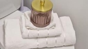A white toilet with a gold lid and a folded white towel on top. - FC Platinum Accommodation (Watford)